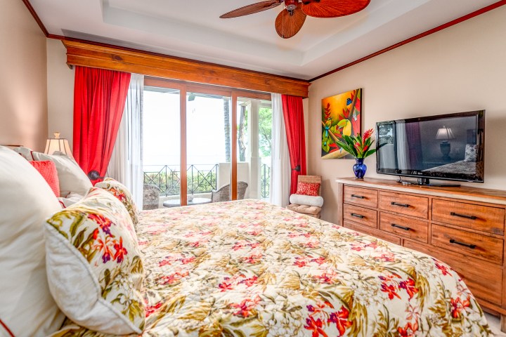 Bedroom with floral bedding, TV on dresser, balcony view, and red curtains.