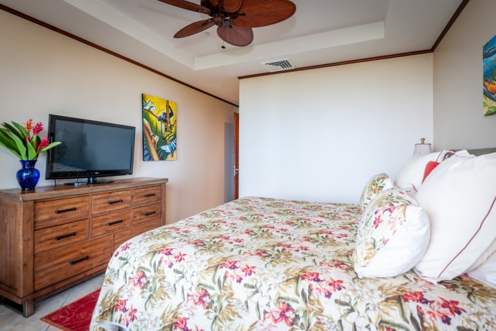 Bedroom with floral bedding, wooden dresser, TV, ceiling fan, and colorful artwork on the wall.