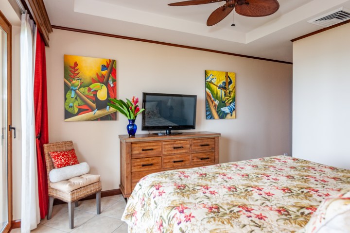 Bedroom with floral bedspread, TV on wooden dresser, and colorful tropical paintings on the wall.