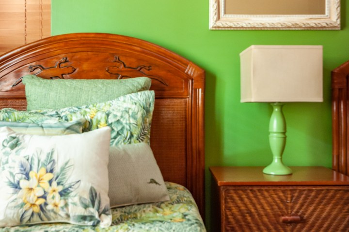 Tropical themed bedroom with green wall, wooden bed, floral bedding, and bedside table with lamp and artwork.