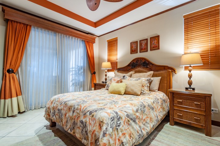 Cozy bedroom with patterned bedspread, orange curtains, and two bedside lamps.