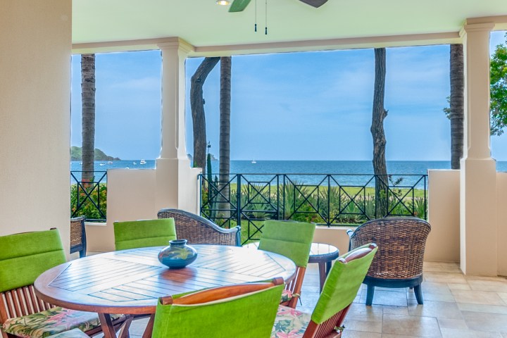 Covered patio with round wooden table, green chairs, and ocean view.