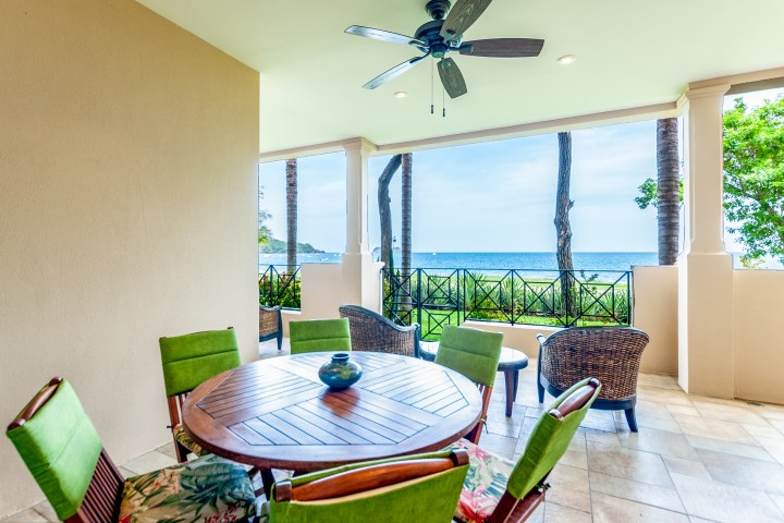 Covered patio with table, chairs, ceiling fan, and ocean view through columns.
