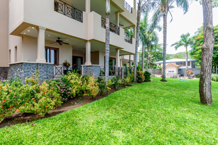 Three-story building with balconies, surrounded by garden and palm trees.