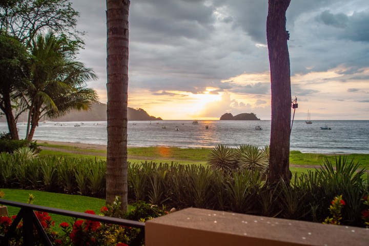 Sunset over a tropical beach with boats on the water and palm trees in the foreground.