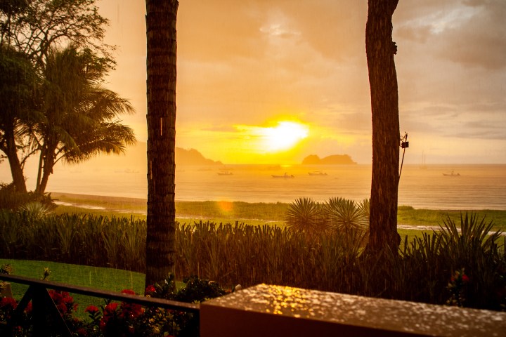 Sunset over a rainy beach with silhouettes of trees and distant boats.