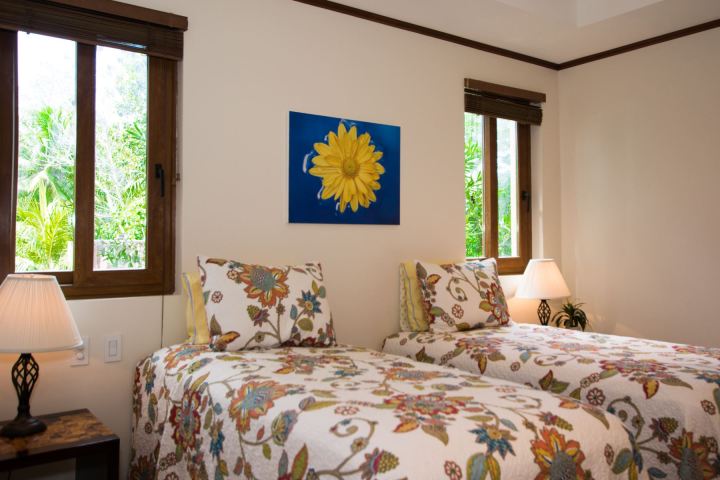 Bedroom with two floral beds, sunflower art, lamps, and two windows with greenery outside.