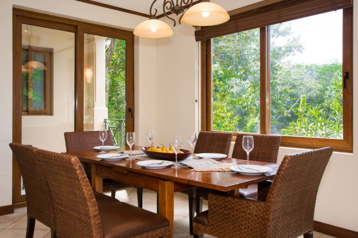 Dining room with six chairs, a wooden table set with plates and glasses, and large windows with garden view.