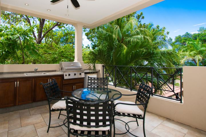Outdoor patio with grill, table, chairs, and lush green trees in the background.