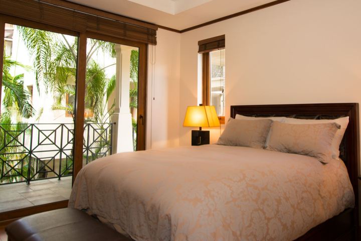 Cozy bedroom with a large bed, lamp, and window view of tropical plants on a balcony.