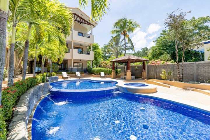 Tropical resort pool with palm trees, lounge chairs, and gazebo near a three-story building.