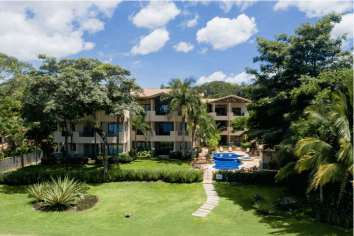 Lush garden with a path leading to a modern building and pool on a sunny day.