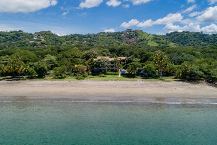 Beachfront view with palm trees, clear water, and hills with houses in the background.