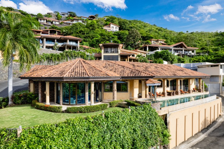 Luxury hillside villa with terracotta roof, pool, and lush greenery under a blue sky.