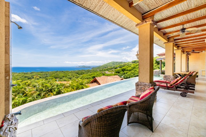 Covered patio with wicker chairs overlooking ocean and lush hills.