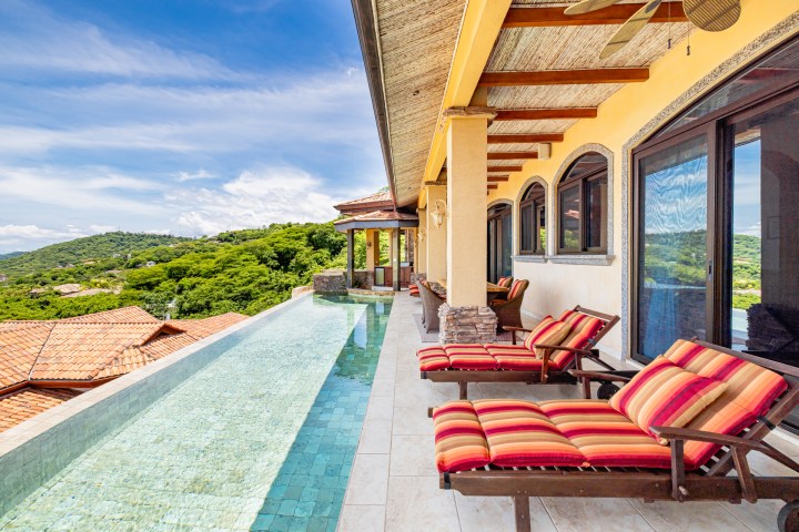 Infinity pool and sun loungers on villa terrace with scenic view of hills.