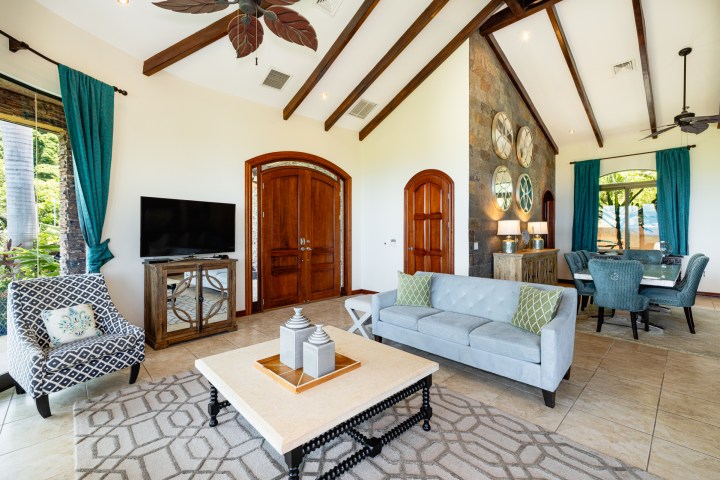 Living room with vaulted ceiling, blue sofa, patterned chair, TV, dining area, and large wooden doors.