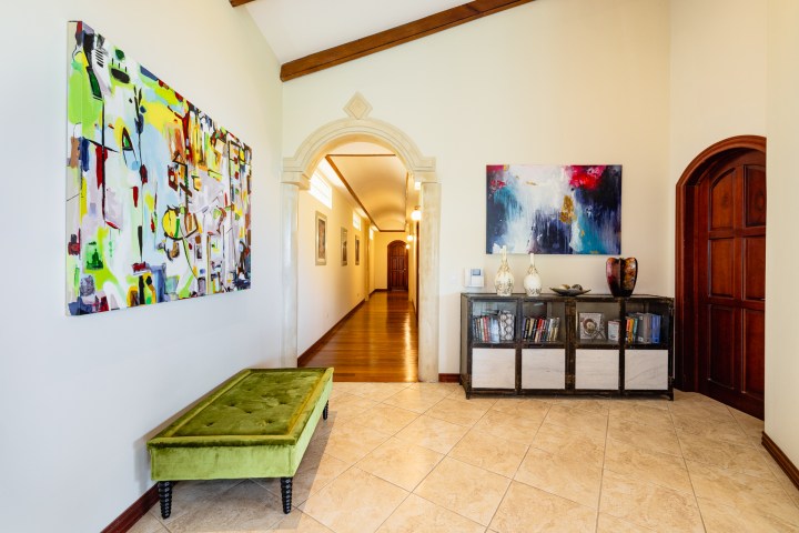 Hallway with archway, colorful abstract paintings, green bench, and a bookshelf with vases.
