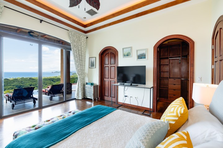Room with bed, TV, and open balcony doors showing ocean view and chairs outside.