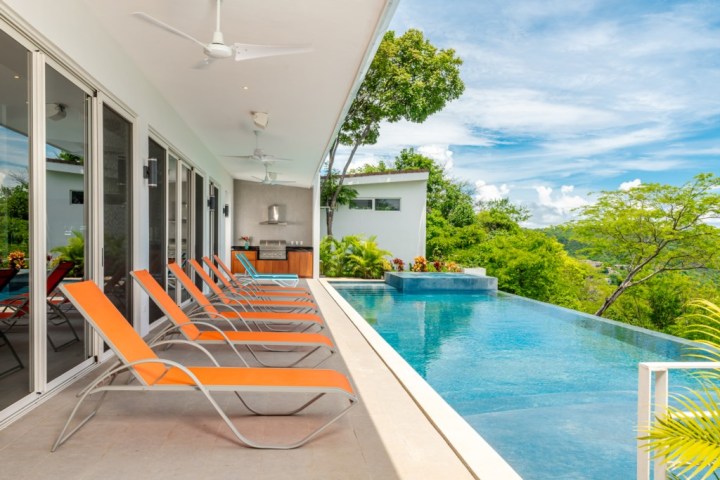 Modern patio with orange lounge chairs beside a sleek infinity pool, overlooking lush greenery and sky.
