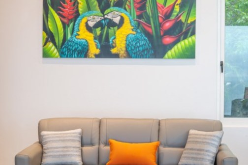 Living room with gray sofa, colorful parrot artwork, round wood coffee table, and window.