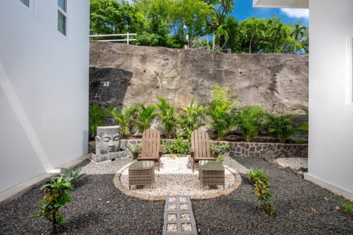 Outdoor garden with two wooden chairs, a Buddha sculpture, tropical plants, and a stone pathway.