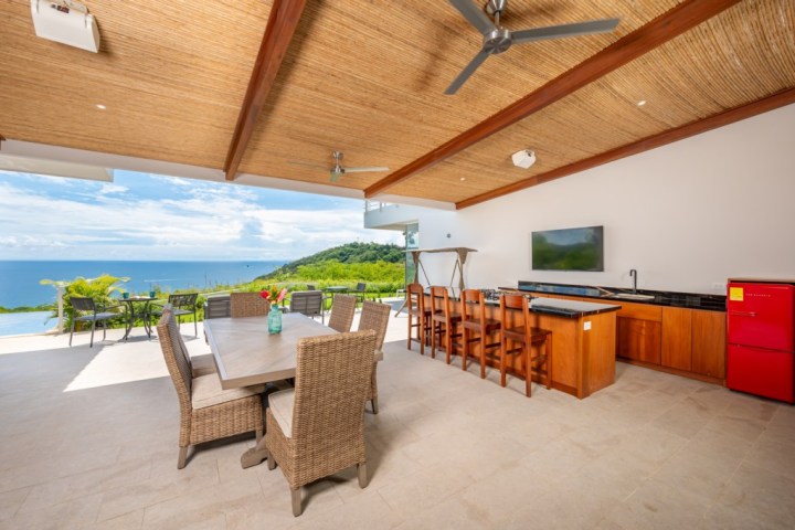 Open-air kitchen and dining area with ocean view, bar, and wicker chairs.