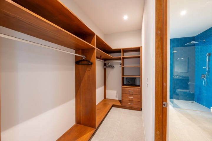 Wooden walk-in closet with open shelves and hangers, next to a bathroom with blue tiles and a shower.