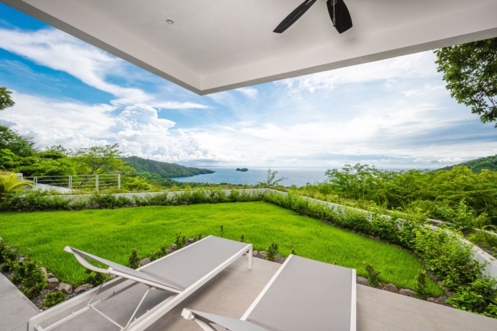 Terrace with loungers overlooking a lush green garden and ocean view under a blue sky.