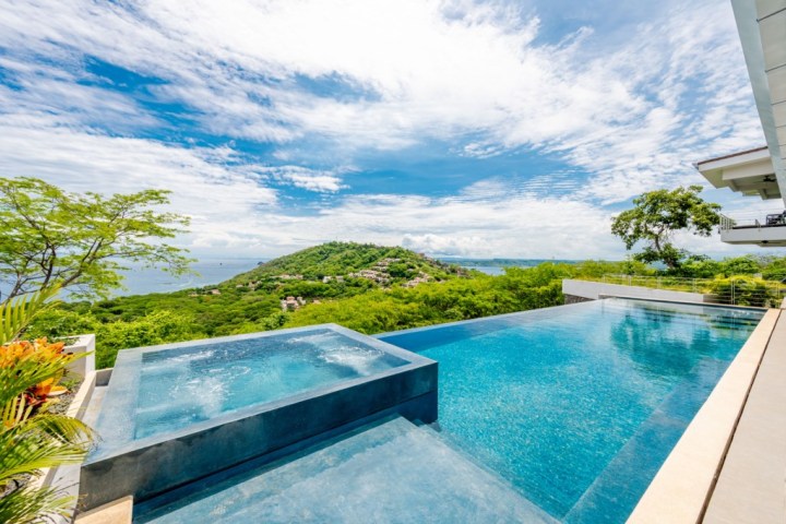 Infinity pool overlooking a lush green hill and ocean under a blue sky with clouds.