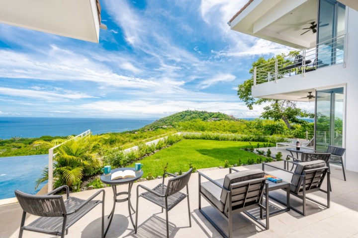 Modern patio with chairs facing ocean view and grassy hill under blue sky.