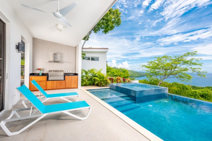 Outdoor patio with two blue loungers, grill, and infinity pool overlooking ocean and trees.