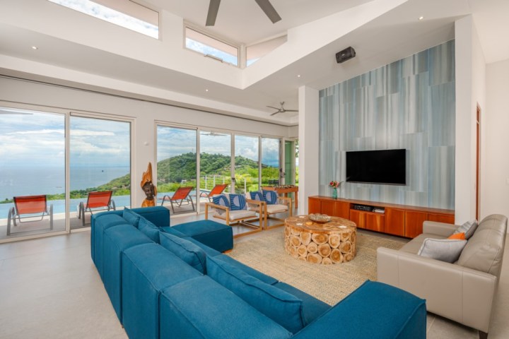 Modern living room with ocean view, blue couches, wall TV, and round wooden table.