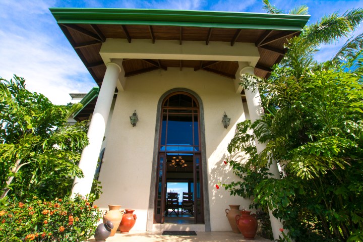 Entrance of a house with white columns and greenery around, featuring large windows.