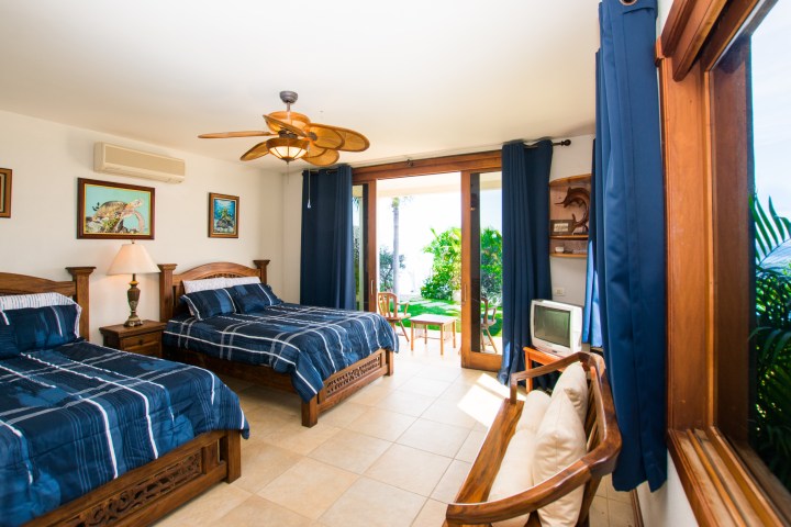 Bedroom with two beds, wooden furniture, ceiling fan, and view of garden through sliding glass door.