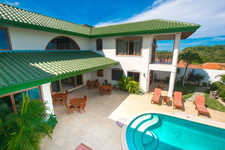 Courtyard view of a two-story house with a green roof, pool, loungers, and patio tables.
