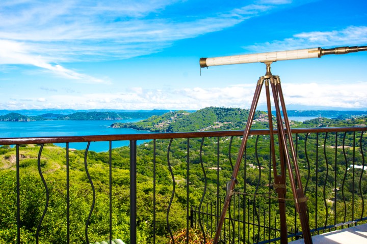 A telescope on a balcony overlooks a lush green landscape and a blue ocean under a clear blue sky.