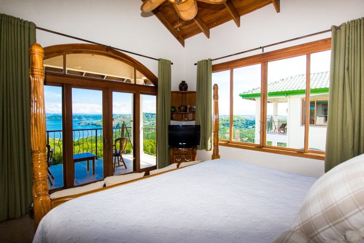 Bedroom with large windows, balcony view of lake and hills, wooden bed frame, and ceiling fan.