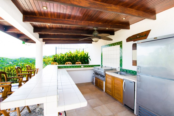 Outdoor kitchen with tile countertops, wooden chairs, and scenic view in the background.
