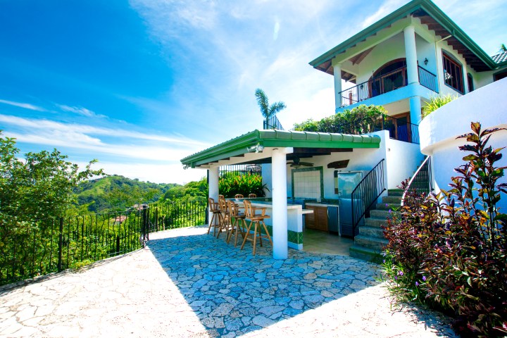 Outdoor patio with a bar area overlooking lush green landscape and distant hills under a clear blue sky.