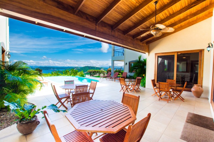 Outdoor patio with wooden tables and chairs overlooking a pool and ocean under a wooden ceiling.