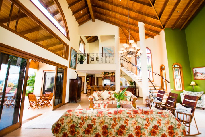 Spacious living room with high wooden ceiling, floral furniture, staircase, and large windows.