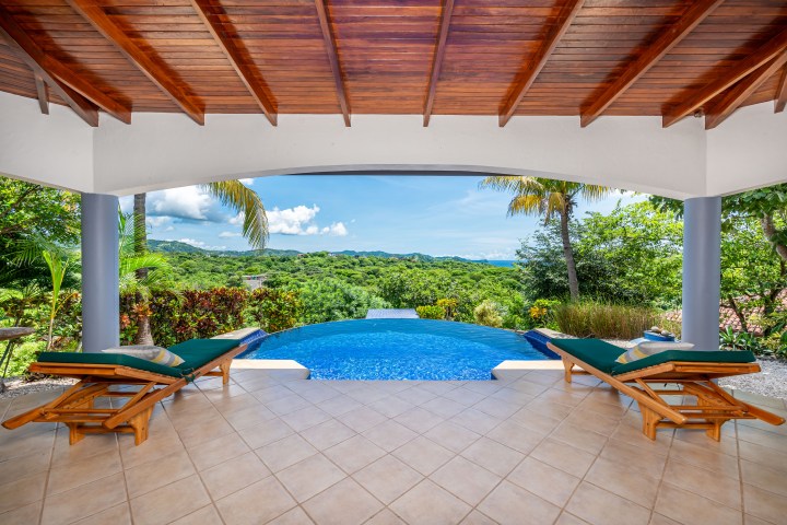 Covered patio with loungers overlooking a pool and lush green landscape.
