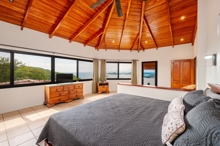 Bedroom with wooden ceiling, large windows, ocean view, and modern furnishings.