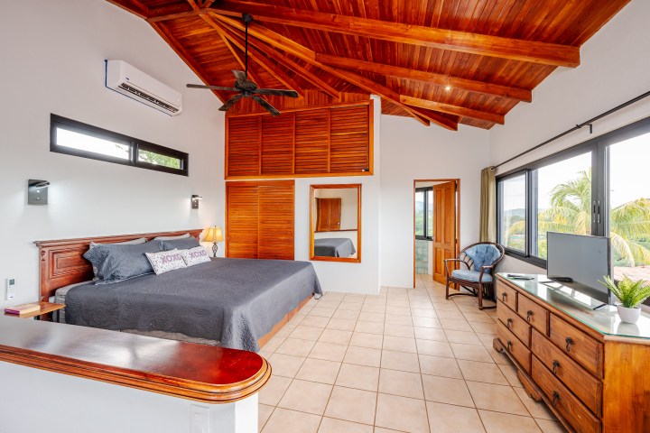 Bedroom with wooden ceiling, king bed, dresser, TV, armchair, large window, and plant