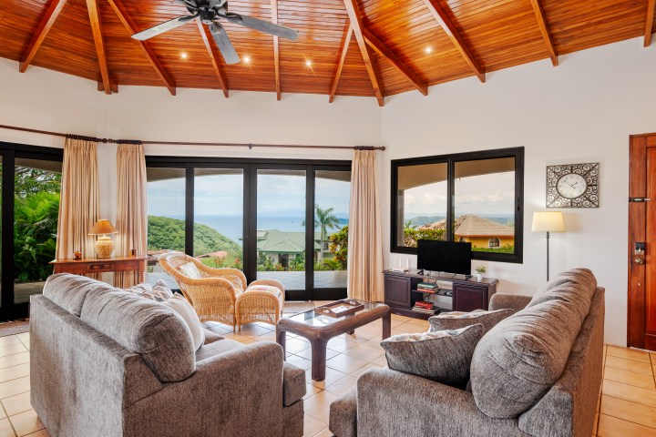 Cozy living room with sofas, wicker chairs, TV, and sea view through large windows; wooden ceiling and tiled floor.