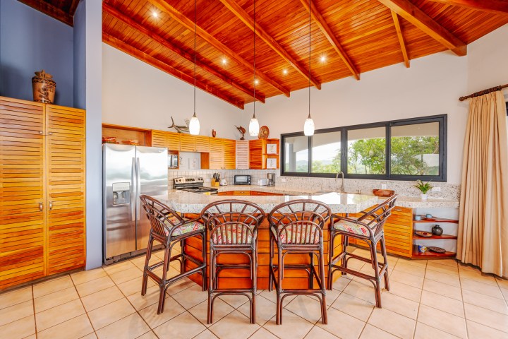 Spacious kitchen with wooden ceiling, bar stools, large counter, and stainless steel fridge.