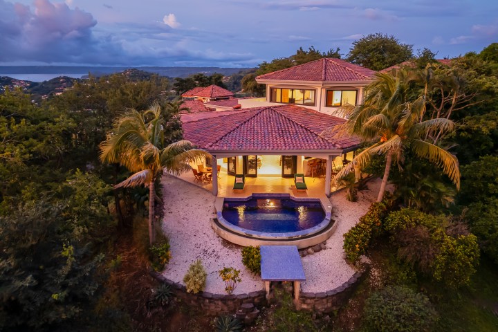 Aerial view of a villa with a pool, surrounded by palm trees at dusk.