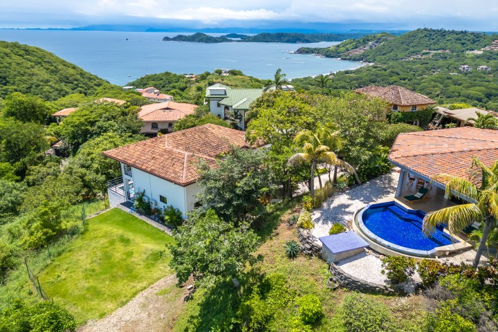 Aerial view of hillside homes with terracotta roofs, pool, and ocean in the background.