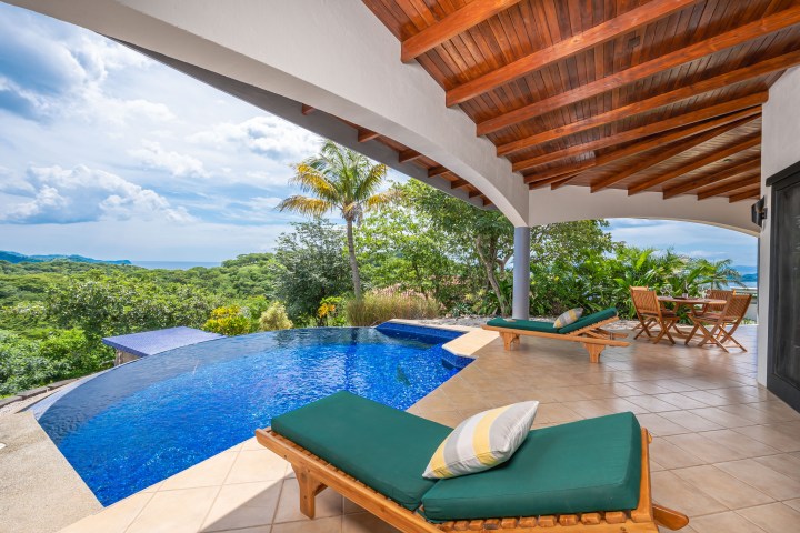 Covered patio with pool, lounge chairs, table, and lush greenery overlooking a scenic view.